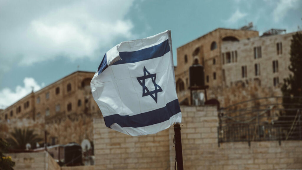 Un primo piano di una bandiera israeliana, con alcuni edifici sfocati di Gerusalemme sullo sfondo.