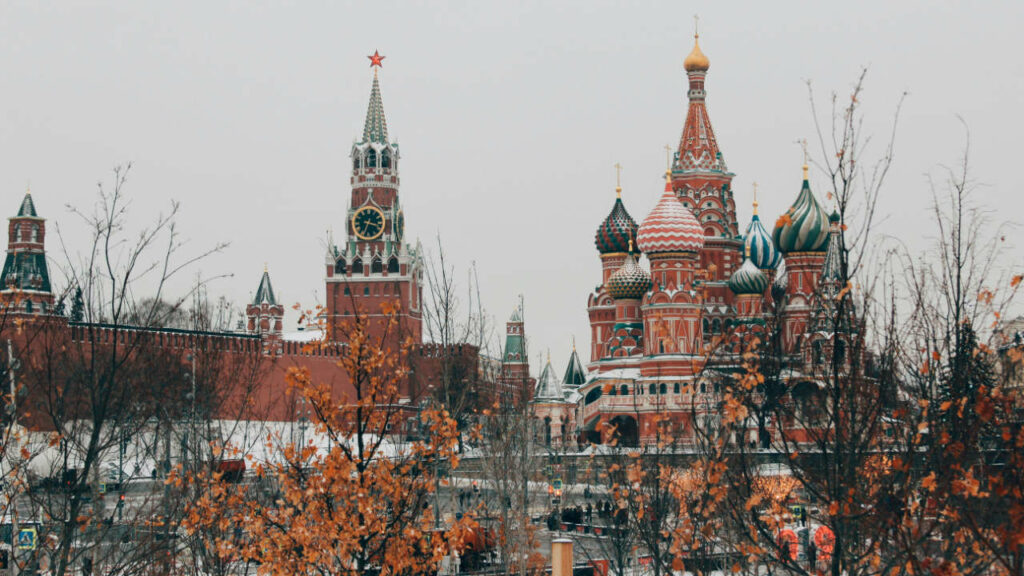 Foto del Cremlino di Mosca, con cielo grigio, e in primo piano alcuni rami con foglie dai colori autunnali ad oscurarne parzialmente la vista.